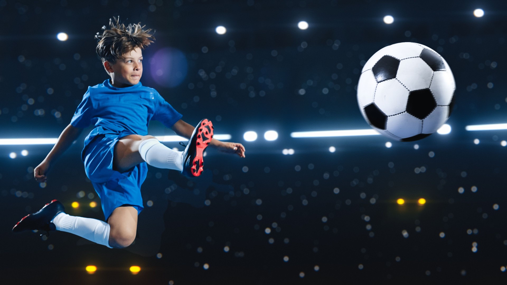 Aesthetic Shot Of Athletic Child Soccer Football Player Jumping And Kicking Ball Mid Air On Black Background Under Spotlight. Young Boy Scoring Beautiful Winning Goal During Championship Final Match